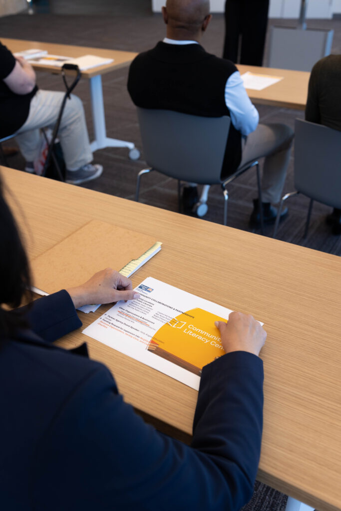 Person reviewing a printed agenda titled "Community Literacy Center" during a meeting, illustrating potential communication barrier through informational materials.
