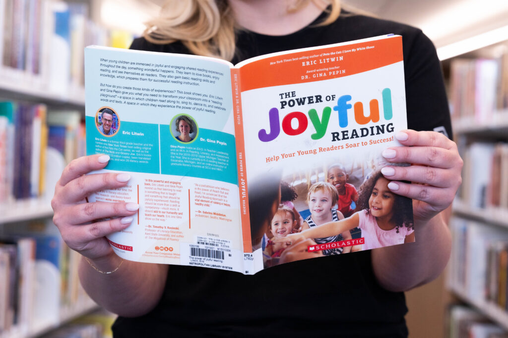 Person holding “The Power of Joyful Reading” book in a library, supporting adult literacy efforts.